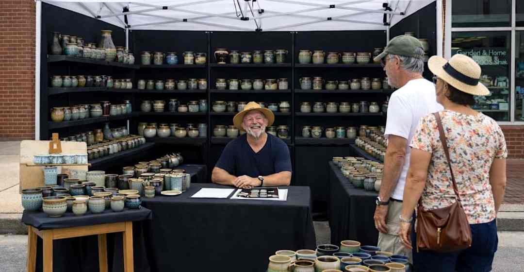 An artist sitting under his popup tent smiling with his handmade pottery displayed.